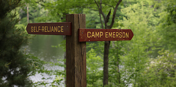 Wooden signpost in a lush green forest by a lake, with two directional signs reading "SELF-RELIANCE" and "CAMP EMERSON".