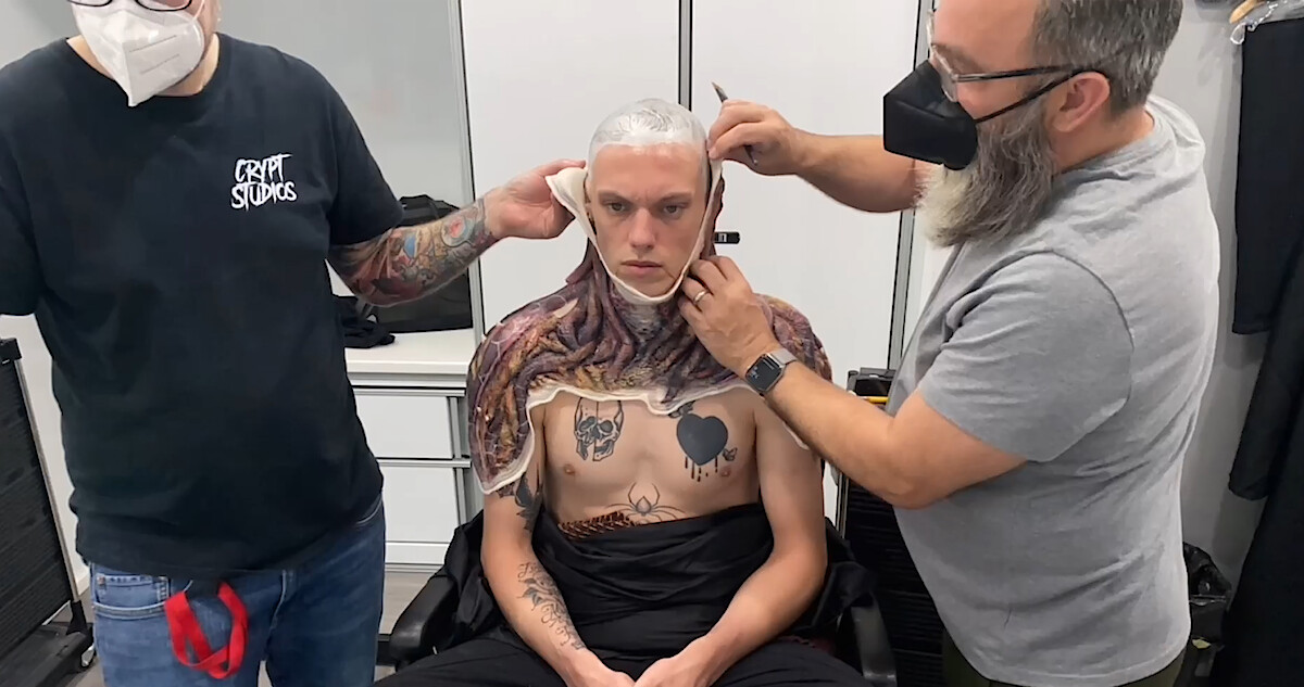 Jamie Campbell Bower sits in a makeup chair while two artists apply a Vecna prosthetic to his head and upper body.