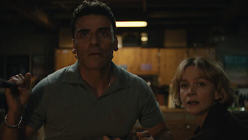 A worried man and woman in a dimly lit basement kitchen, looking intently at something off-screen, with wood cabinets and cluttered shelves in the background.