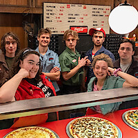 Group of young adults posing behind the counter of a retro pizza shop with three pizzas on display, wearing casual and work uniforms, with wood-paneled walls, a menu board, and warm lighting in the background.