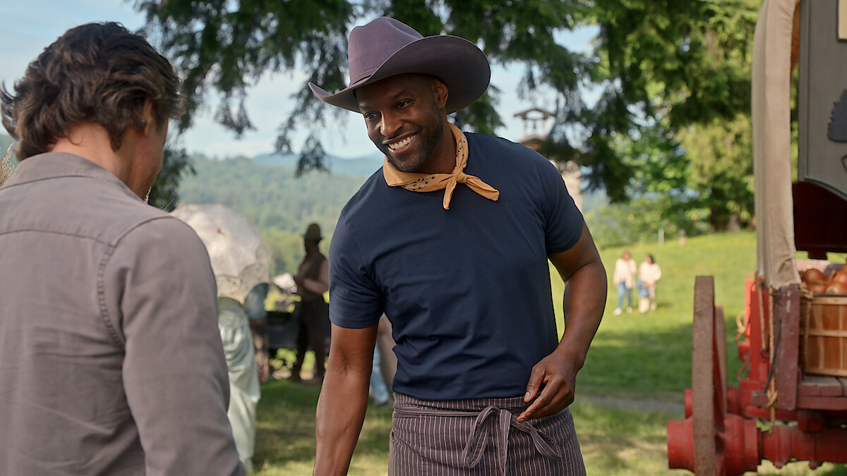 A smiling man in a cowboy hat and neckerchief stands outdoors near a wooden wagon, talking to another person. The setting is a green park or field with trees and people in the background, suggesting a friendly, rustic event.
