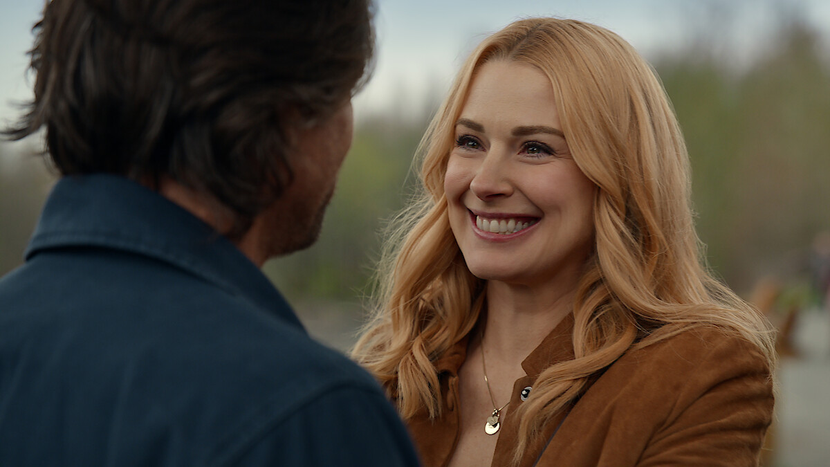 A smiling woman with blonde hair wearing a tan jacket faces a man outdoors, with blurred greenery in the background, suggesting a happy, casual moment between them in a natural setting.
