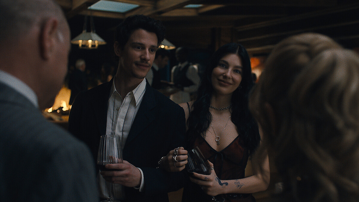 A man and woman dressed formally at a dimly lit indoor social gathering, holding drinks and smiling while conversing with other guests in an elegant setting.