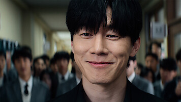 A young man with black hair smiles confidently in a school hallway, surrounded by other students in uniforms, with bright lighting and a lively environment.