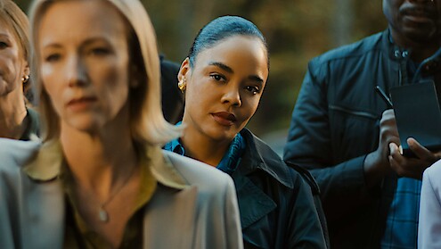 A woman in focus stands among a group of people outdoors, listening attentively, with trees in the background. The scene suggests a serious or formal gathering, possibly a press conference or official announcement.