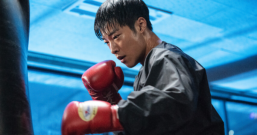 In a blue-lit room, a man wearing a black jacket and red boxing gloves prepares to hit a punching bag.