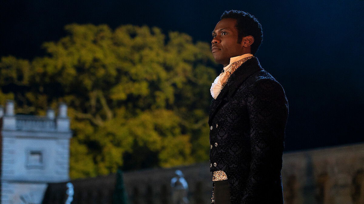 A man in Regency-era formal attire stands outdoors at night, beside a stone building with large trees in the background, lit dramatically.