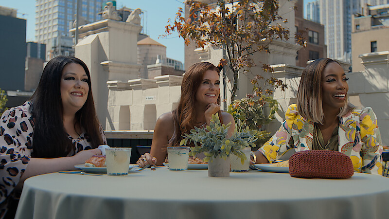Three women sit at an outdoor rooftop table in a city, smiling and enjoying drinks and food, surrounded by buildings and greenery in a casual, urban daytime setting.