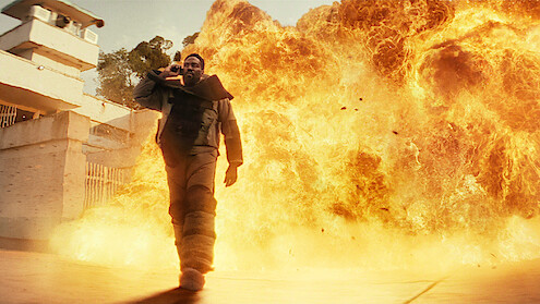 A person in tactical gear walks confidently away from a huge explosion outside a building, surrounded by dramatic flames and intense light in an urban setting.