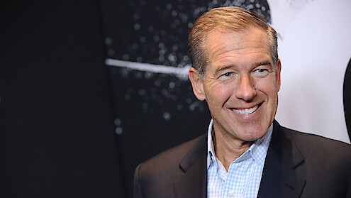 Man in a suit smiling in front of a dark, abstract background with white speckles and lines, possibly at an event or exhibition.
