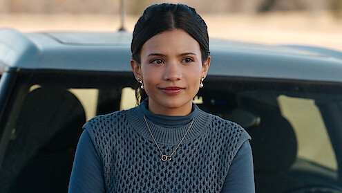 Nikki Rodriguez as Jackie wears a blue sweater and stands by a car in 'My Life with the Walter Boys' Season 2.