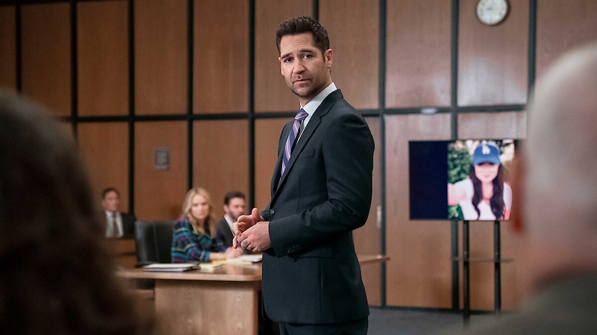 Man in suit stands in courtroom, addressing others. People sit at a table in the background. A digital screen shows a woman’s photo. Wood panel walls and a clock indicate a formal court setting.