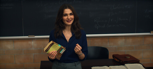Rachel Weisz as The Protagonist standing in front of classroom chalkboard, holding book and smiling, with desk in foreground holding papers and a briefcase, academic setting.