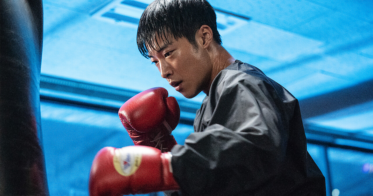 In a blue-lit room, a man wearing a black jacket and red boxing gloves prepares to hit a punching bag.