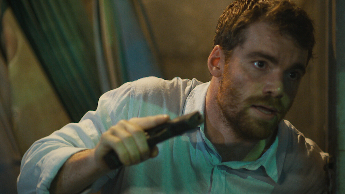 Gabriel Basso as Peter Sutherland leans on a wall holding a gun in Season 2 of 'The Night Agent'