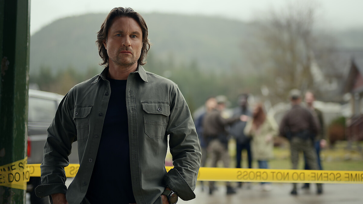 A serious man stands in front of yellow police tape at a crime scene, with law enforcement and bystanders in the background and a misty, forested landscape behind them.