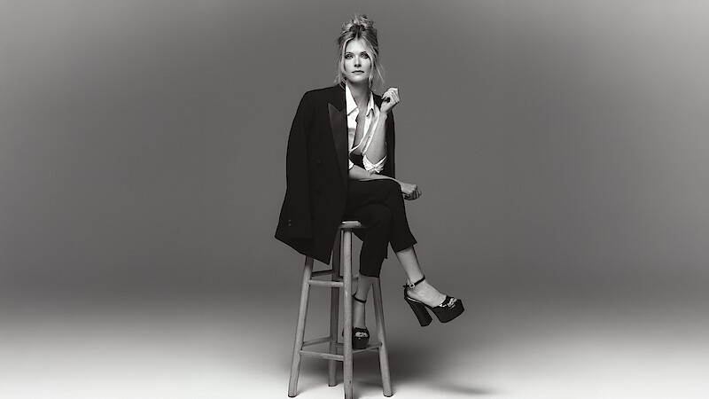 Meghann Fahy sits on a stool in a chic black suit with white button up blouse with legs crossed and platform heels.