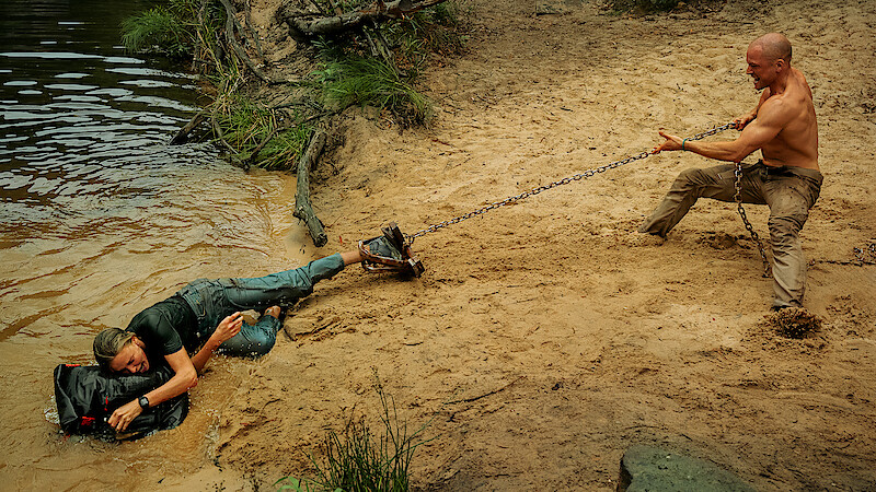 A shirtless man on sandy shore pulls a chain attached to a person partially in water, who is holding a black bag, with trees and water in the natural outdoor background.