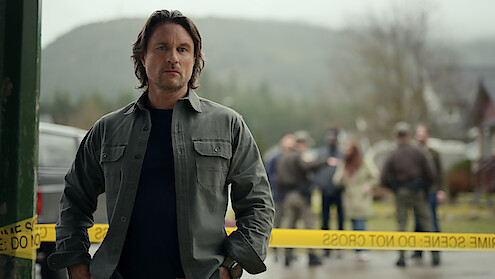 A serious man stands in front of yellow police tape at a crime scene, with law enforcement and bystanders in the background and a misty, forested landscape behind them.