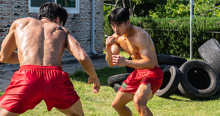 Two muscular  boxers in red shorts square off on a lawn with old tires stacked up behind them. 