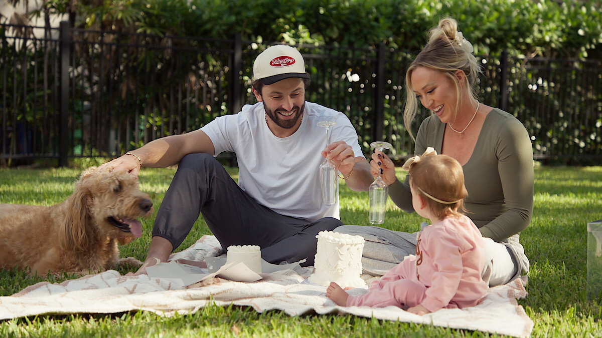 Baker Mayfield with his family and dog enjoying a picnic on a blanket in a sunny backyard, playing with objects and smiling, surrounded by greenery and a fence in the background.