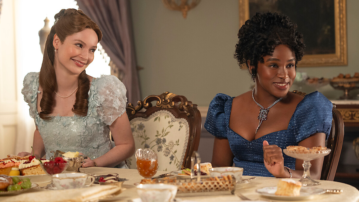Two women in elegant dresses sit at a lavish dining table set with desserts and tea in a well-lit, ornate room with classical decor, enjoying conversation.