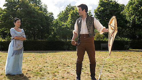 A woman in a light blue dress and a man in period costume with a kite stand on a grassy field in a garden, surrounded by green trees under a partly cloudy sky.