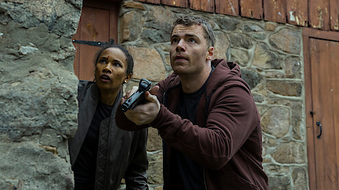 Two people crouch by a stone wall, looking alert and cautious. One holds a gun, both appear focused and ready for action. The setting is rustic, with stone and wood walls suggesting a rural or remote area.