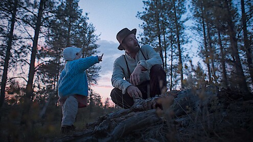 A man in a hat and a child in a blue sweater explore a forest at dusk, surrounded by tall trees. The child points at something, creating a sense of curiosity and peacefulness in the natural setting.