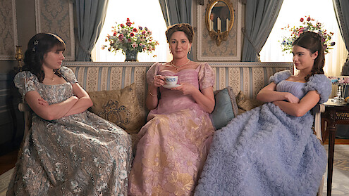 Three women in elegant period dresses sit on a vintage sofa in a richly decorated room, two looking displeased with arms crossed, one smiling with a teacup, creating a formal yet tense atmosphere.