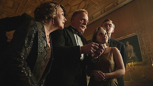 Four well-dressed people in formal evening attire stand close together in an ornate, dimly-lit room with a decorated ceiling and classical artwork, appearing to discuss something intently.
