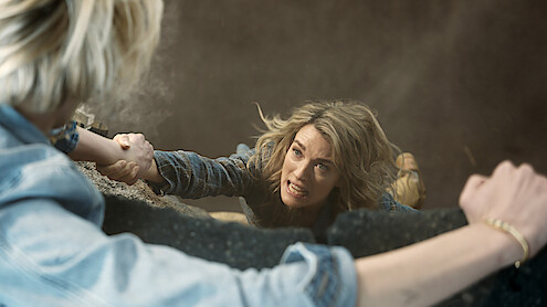 A woman hanging from a ledge, gripping another person's arm, with a distressed expression, surrounded by dust and debris in a tense, dramatic rescue scene.