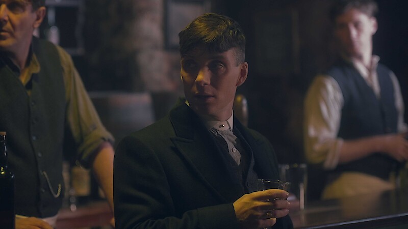 A man holds a drink in a pub with two bartenders behind him. 