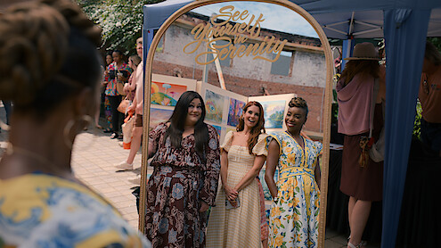 Three women smiling and posing together in front of a decorative mirror at an outdoor event, with other people and a tent in the background. The mirror has the words "See Yourself in Serenity" written on it.