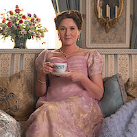 A woman in an elegant period dress sits in a wooden chair in a richly decorated room with bookshelves and a fireplace, appearing calm and contemplative. The atmosphere is refined and historical.