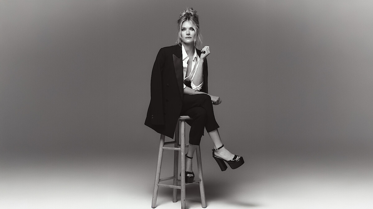 Meghann Fahy sits on a stool in a chic black suit with white button up blouse with legs crossed and platform heels.