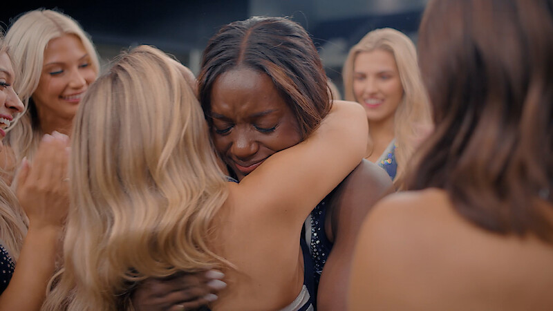A group of women in a celebratory indoor setting, with one woman hugging another who appears emotional while others smile and clap around them.