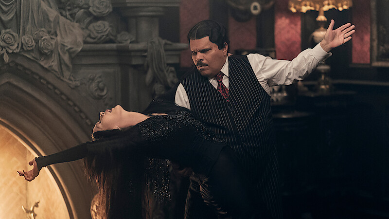 Catherine Zeta-Jones as Morticia Addams and Luis Guzmán as Gomez Addams dancing together in front of a fireplace in 'Wednesday' Season 2.