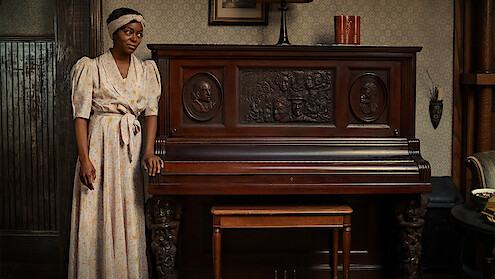 Berniece Charles (Danielle Deadwyler) stands in a white dress at an intricately carved wooden piano. 
