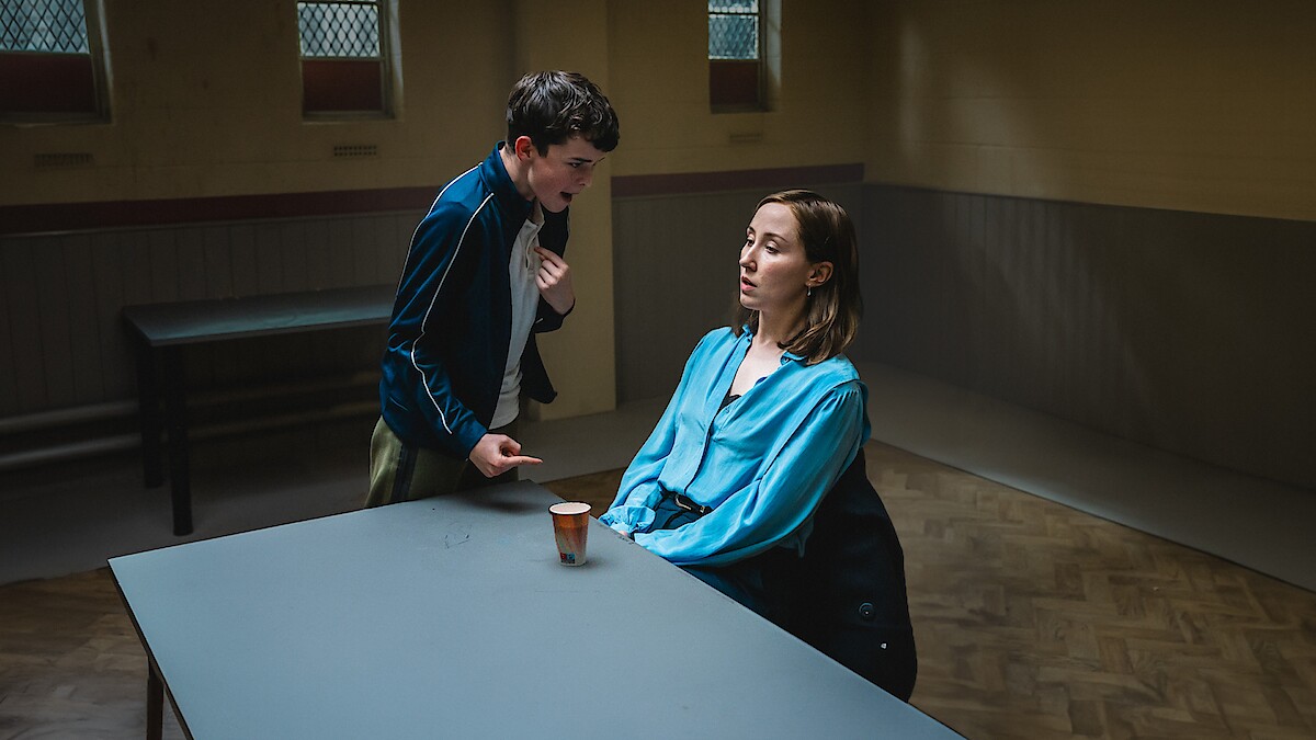 A tense scene shows a young man standing and gesturing intensely at a seated woman in a blue blouse across a table in a dimly lit room with barred windows.