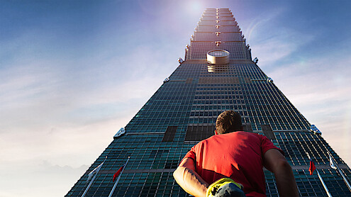 Alex Honnold in a red shirt stands at the base of a tall skyscraper, looking up towards the top under a clear blue sky, suggesting ambition or challenge in an urban environment.