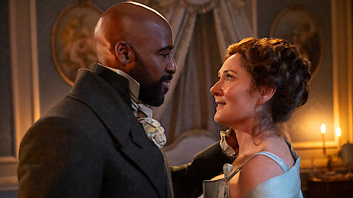 A man and woman in historical clothing gaze at each other in an elegantly decorated, candlelit room with ornate walls and portraits, suggesting a romantic or emotional moment in a period drama setting.