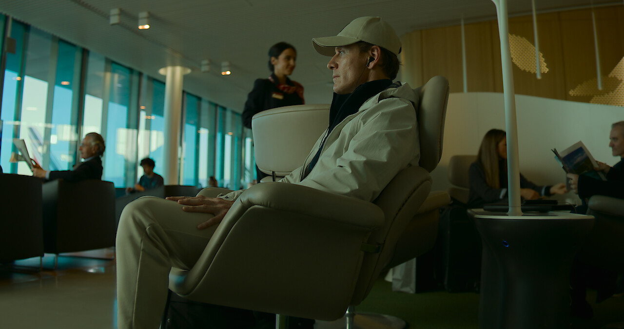 Michael Fassbender sits in a chair wearing a beige baseball hat and jacket in a still from 'The Killer.'
