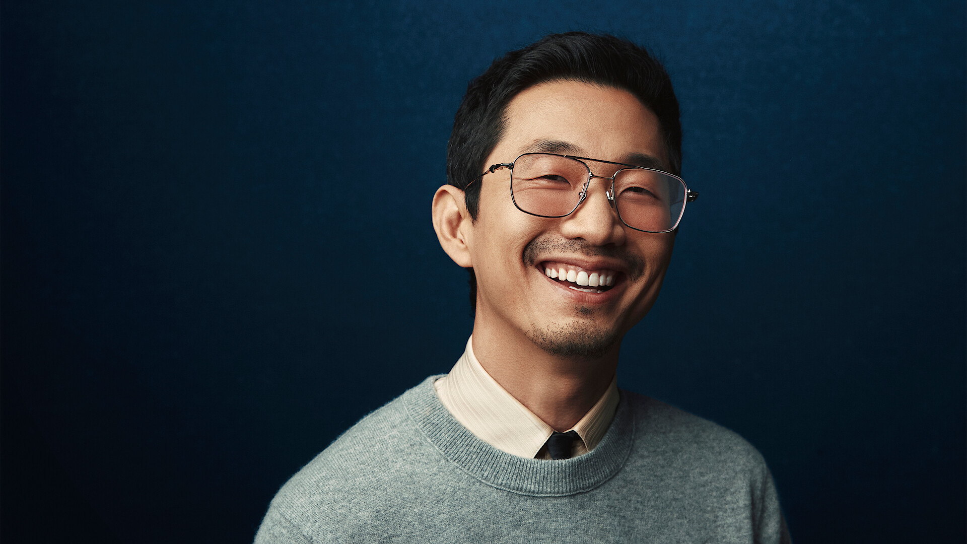 Lee Sung Jin stands in front of a backdrop that shifts from black to dark blue, smiling and looking to camera.