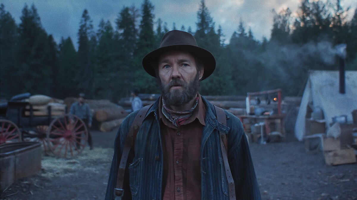 Bearded man in old-fashioned clothing stands in a rustic outdoor camp with wooden wagons, tents, and pine trees, creating a somber and rugged frontier atmosphere at dusk.