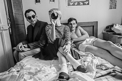 Jean-Luc Godard (Guillaume Marbeck), Jean Seberg (Zoey Deutch), and Jean-Paul Belmondo (Aubry Dullin) sit on a bed in a hotel room as they film Breathless in Richard Linklater's Nouvelle Vague portraying the making of the revolutionary film. 