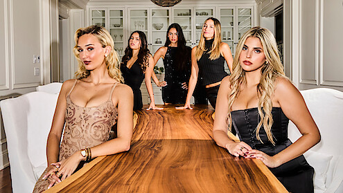 Suede, Emma, Nicole, Emilie, and Jemma in stylish dresses pose around a polished wooden table in a modern dining room with white chairs and cabinets, under warm lighting.