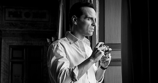 Black and white photo of Andrew Scott as Tom Ripley holding a camera while looking out of a window in Season 1 of 'Ripley'