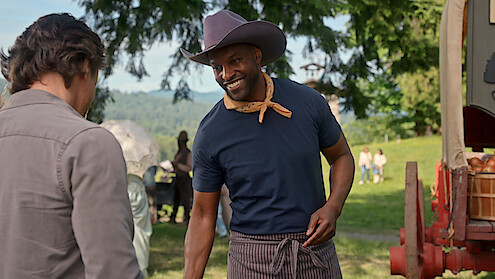 A smiling man in a cowboy hat and neckerchief stands outdoors near a wooden wagon, talking to another person. The setting is a green park or field with trees and people in the background, suggesting a friendly, rustic event.