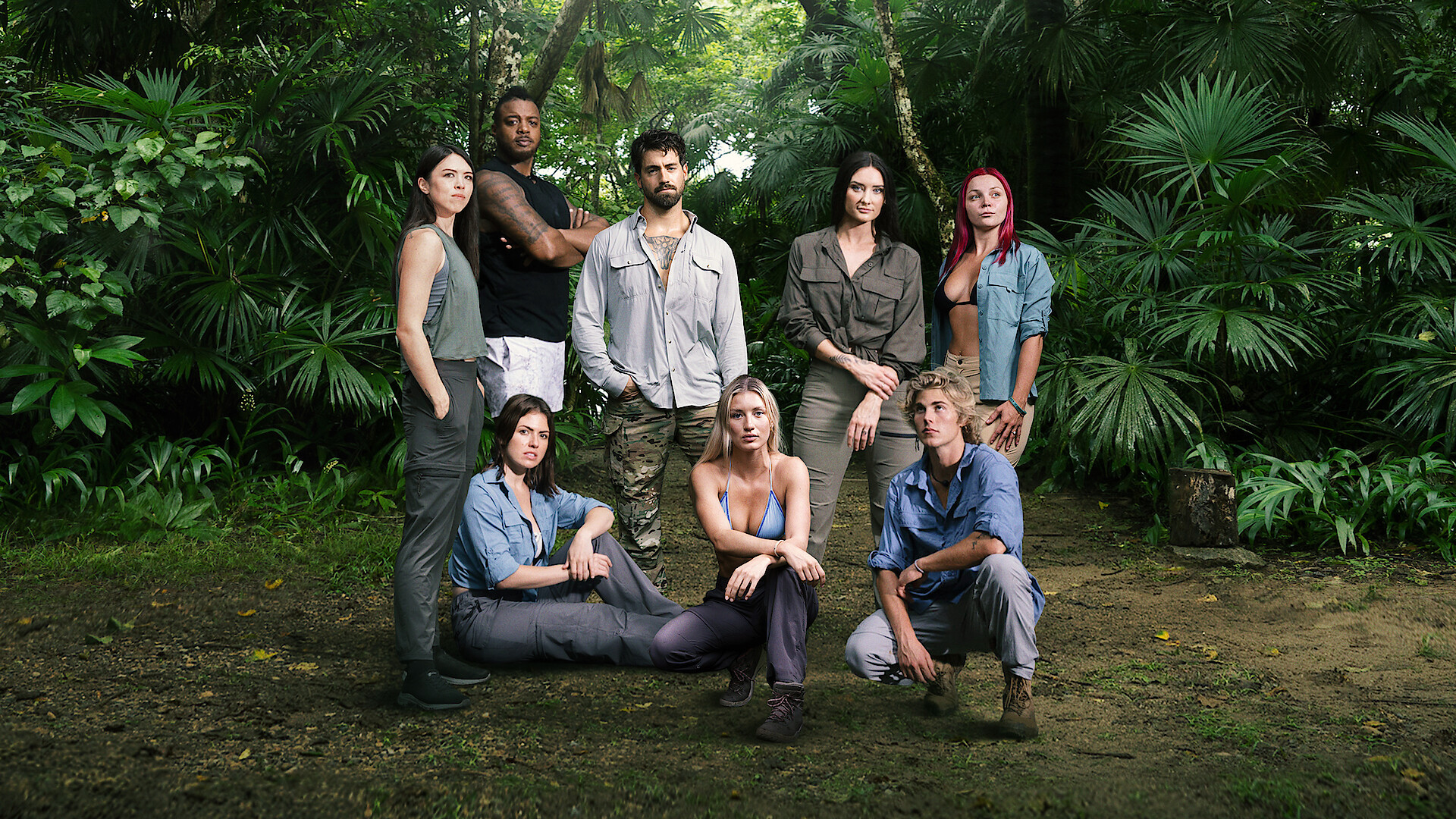Group of eight people posing in a jungle setting, surrounded by lush green foliage, wearing outdoor adventure clothing, with some standing and others sitting on the ground, creating a team or expedition atmosphere.